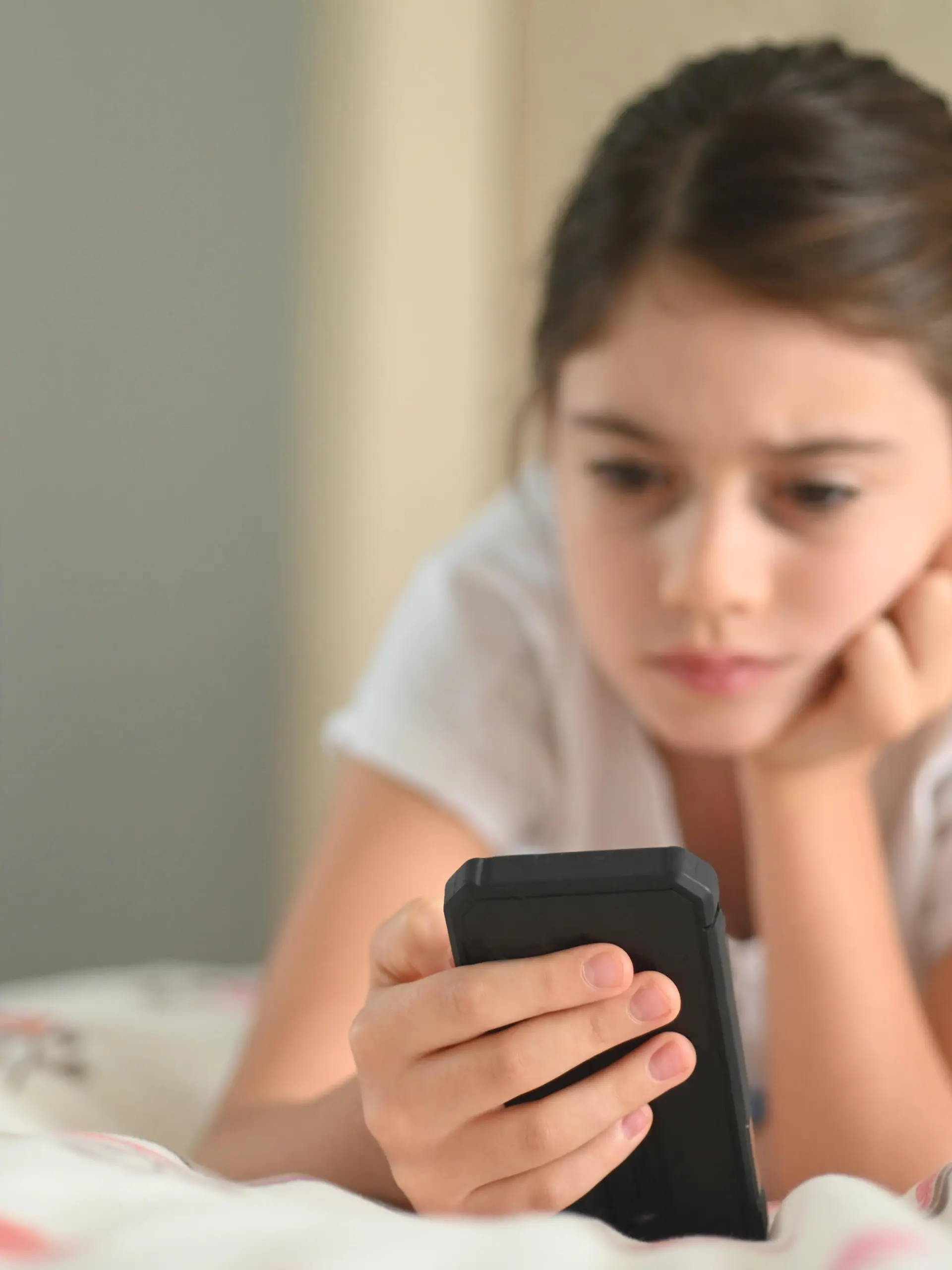 Ein junges Mädchen sitzt auf dem Bett und hält ihr Handy in der Hand. Ihrem Gesichtsausdruck nach scheint sie bedrückt und etwas gesehen zu haben, was ihr nicht gefällt.