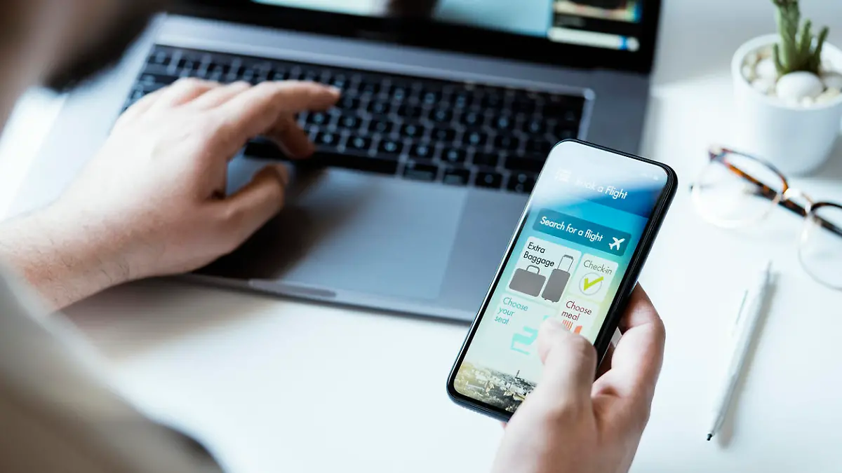 Book a flight, air travel app on smart phone screen, home interior. Hand holding phone, desktop shot.