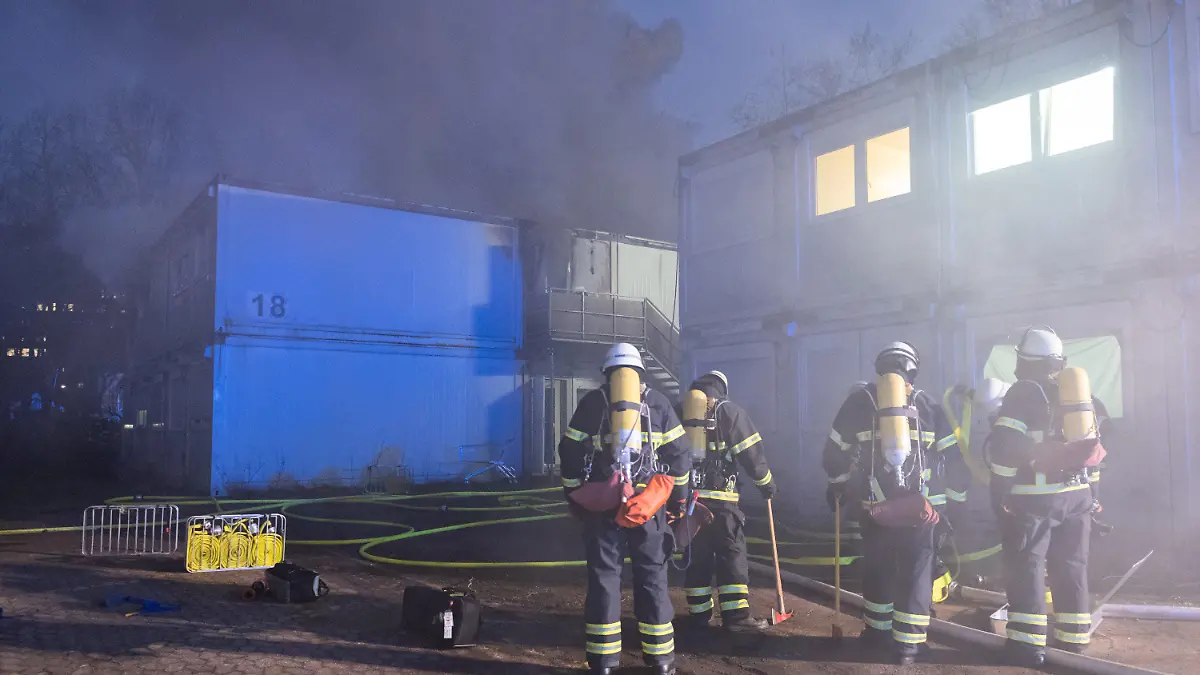 13.02.2023, Hamburg: Feuerwehrleute stehen bei Löscharbeiten vor den verqualmten und teils brennenden Container-Einheiten einer Flüchtlingsunterkunft in Hamburg-Winterhude. Bei dem Brand kam ein Mensch ums Leben. Foto: Jonas Walzberg/dpa +++ dpa-Bildfunk +++