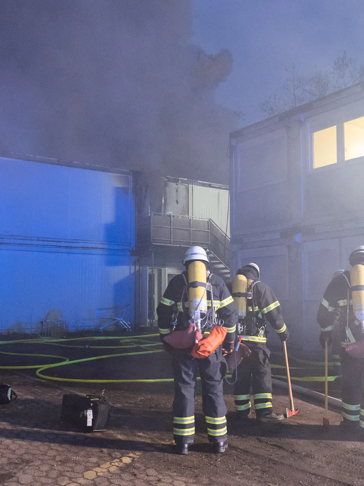 13.02.2023, Hamburg: Feuerwehrleute stehen bei Löscharbeiten vor den verqualmten und teils brennenden Container-Einheiten einer Flüchtlingsunterkunft in Hamburg-Winterhude. Bei dem Brand kam ein Mensch ums Leben. Foto: Jonas Walzberg/dpa +++ dpa-Bildfunk +++