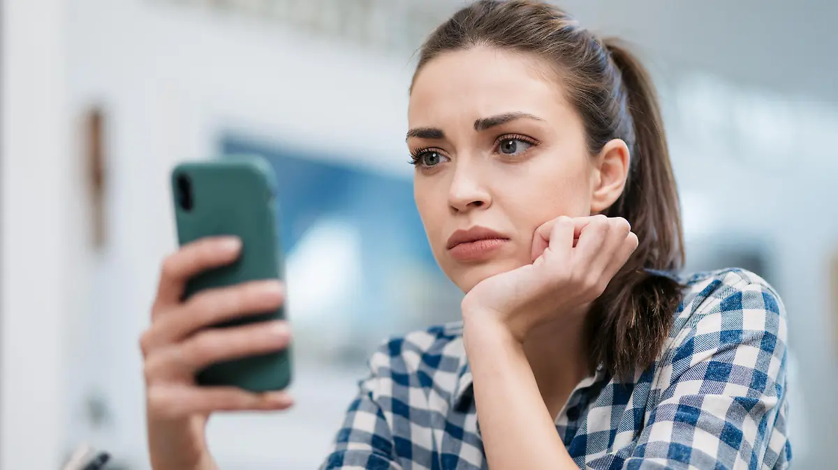 Close up of a depressed Caucasian young woman sitting at home, reading some bad news on her smart phone, feeling sad and worried