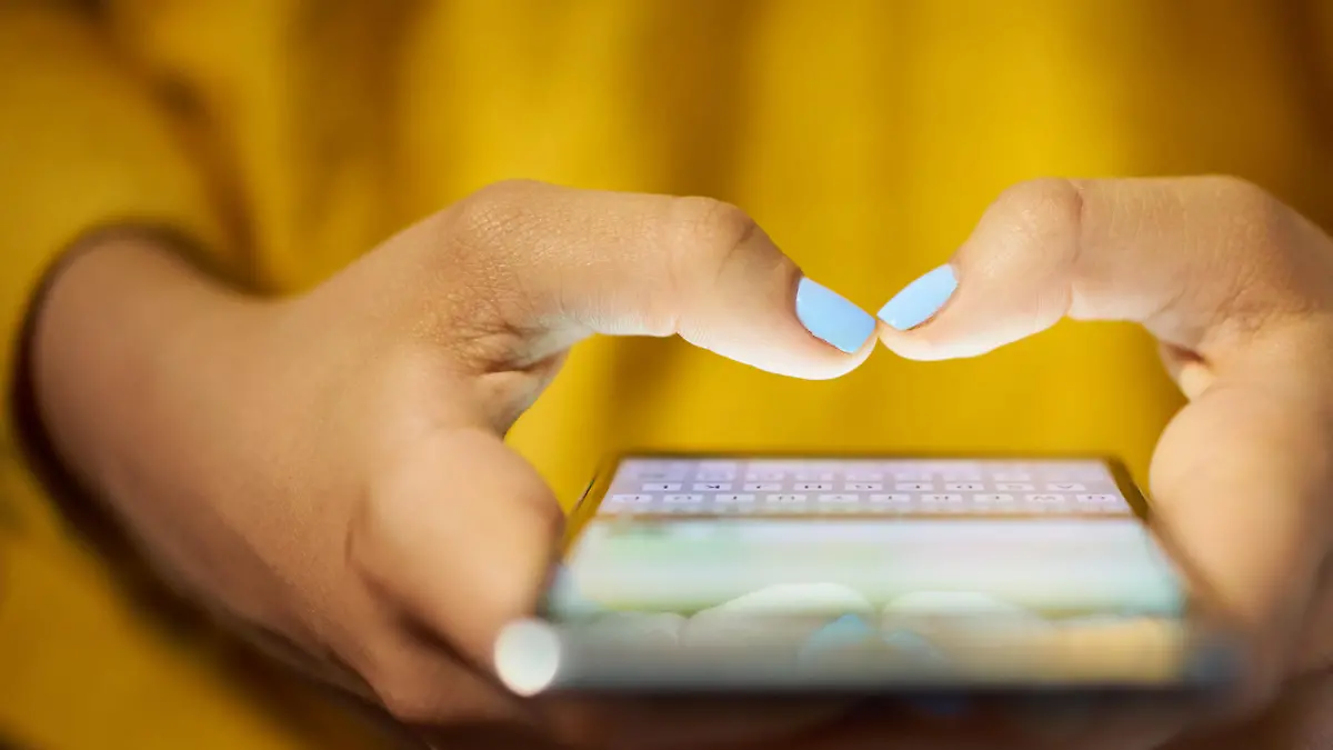 Young woman using cell phone to send text message on social network at night. Closeup of hands with computer laptop in background