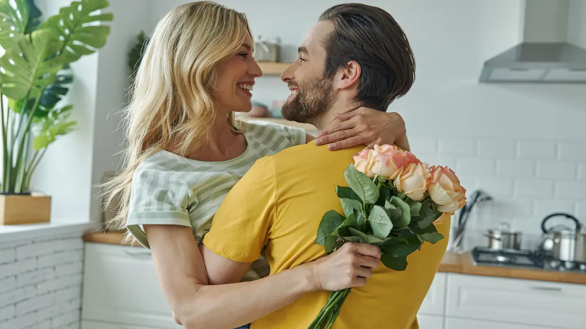 Cheerful young man surprises his girlfriend with bunch of flowers at the kitchen