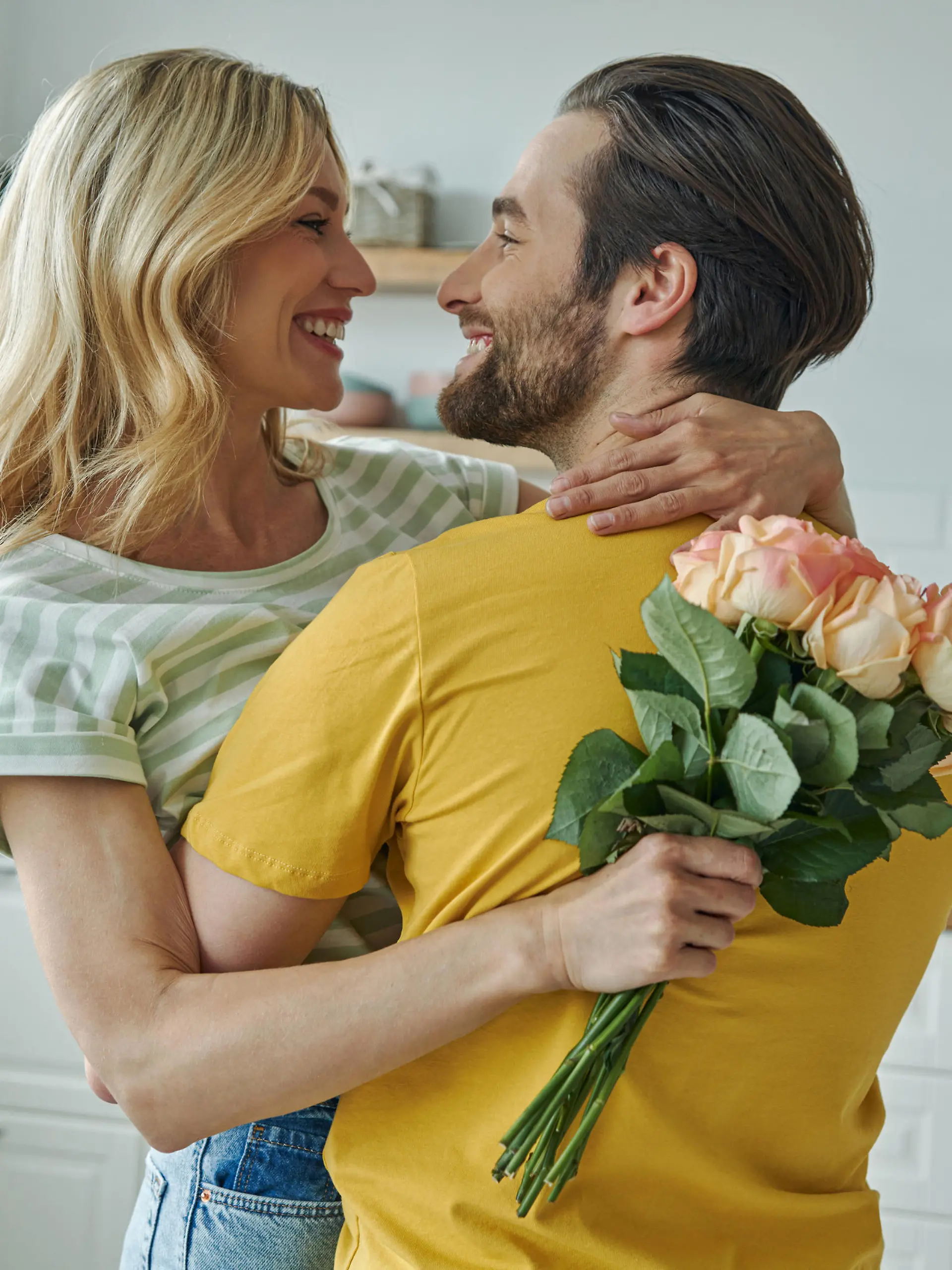 Cheerful young man surprises his girlfriend with bunch of flowers at the kitchen
