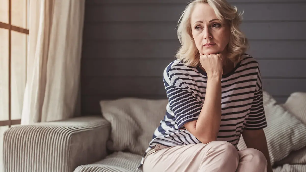 Beautiful sad senior woman is leaning on her hand and looking downward while sitting on couch at home