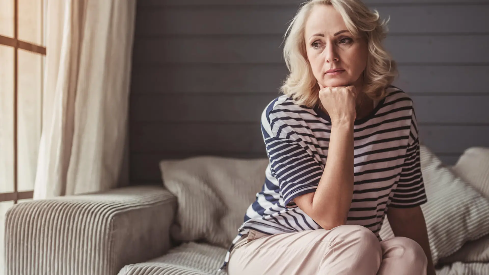 Beautiful sad senior woman is leaning on her hand and looking downward while sitting on couch at home