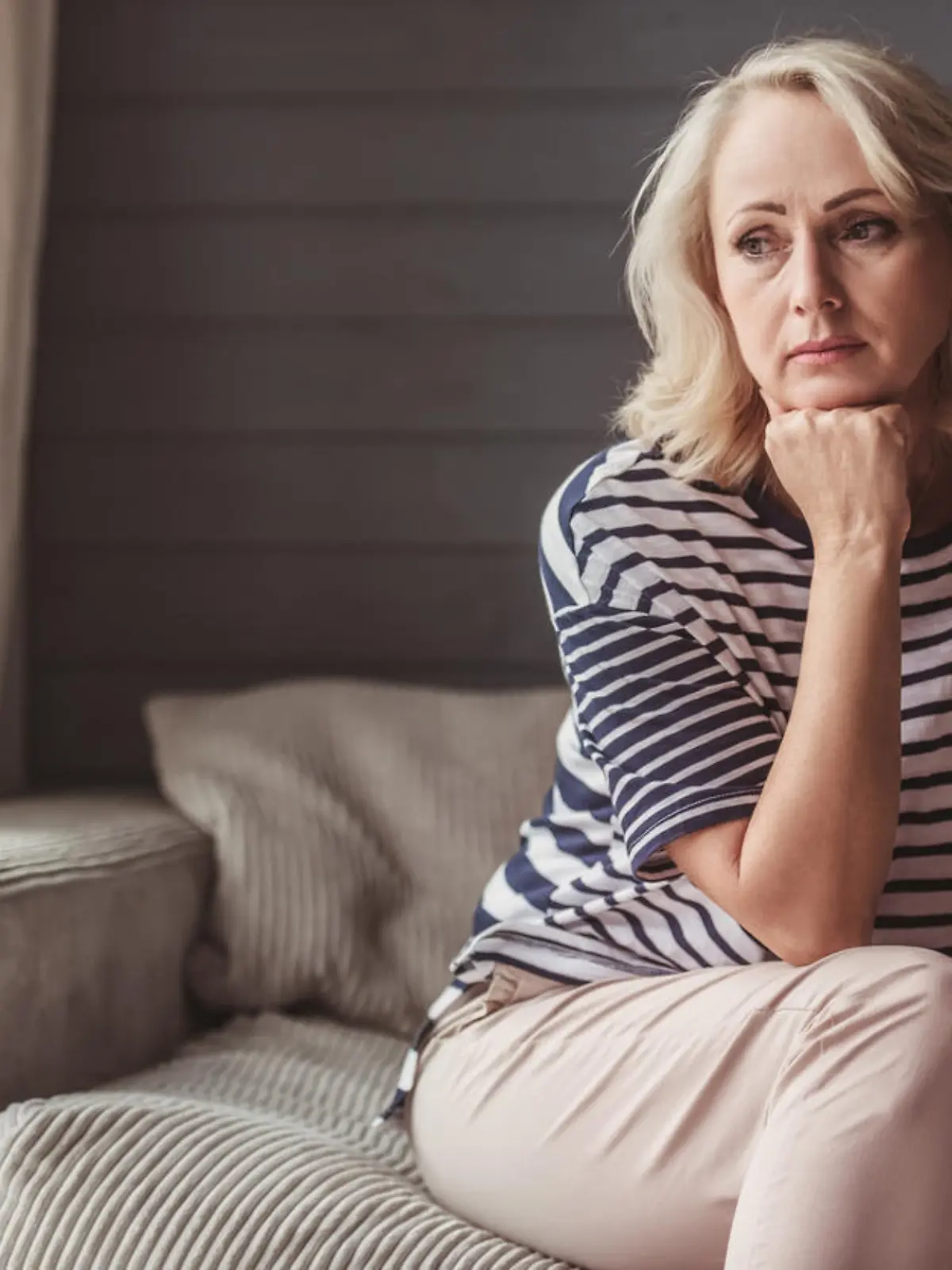 Beautiful sad senior woman is leaning on her hand and looking downward while sitting on couch at home