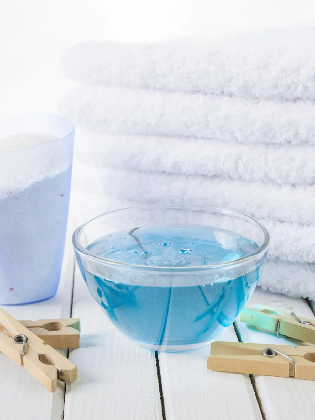 Stack of three white fluffy bath towels, washing powder in measuring cup, blue fabric softener in a glass bowl and wooden clothespins on the background of white boards