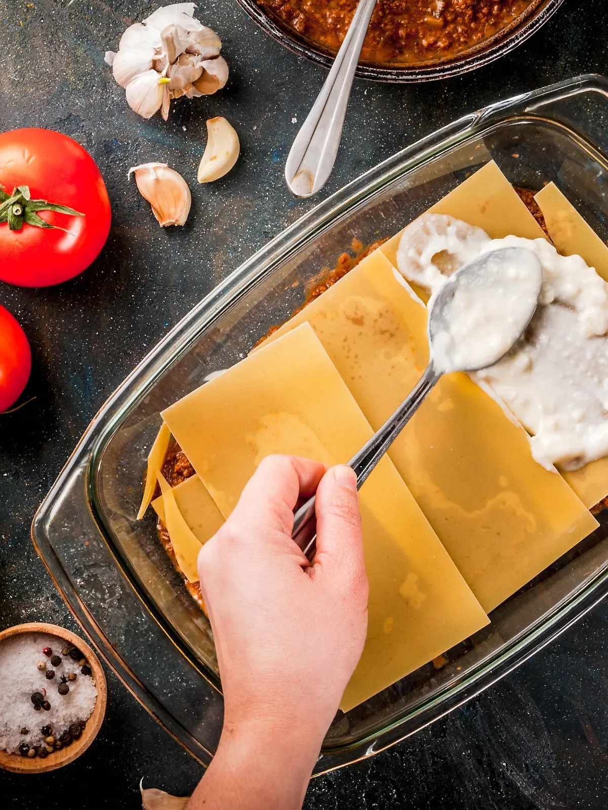 Woman cooking homemade classic lasagna bolognese, on dark blue table; with ingredients, top view copy space, hands in picture