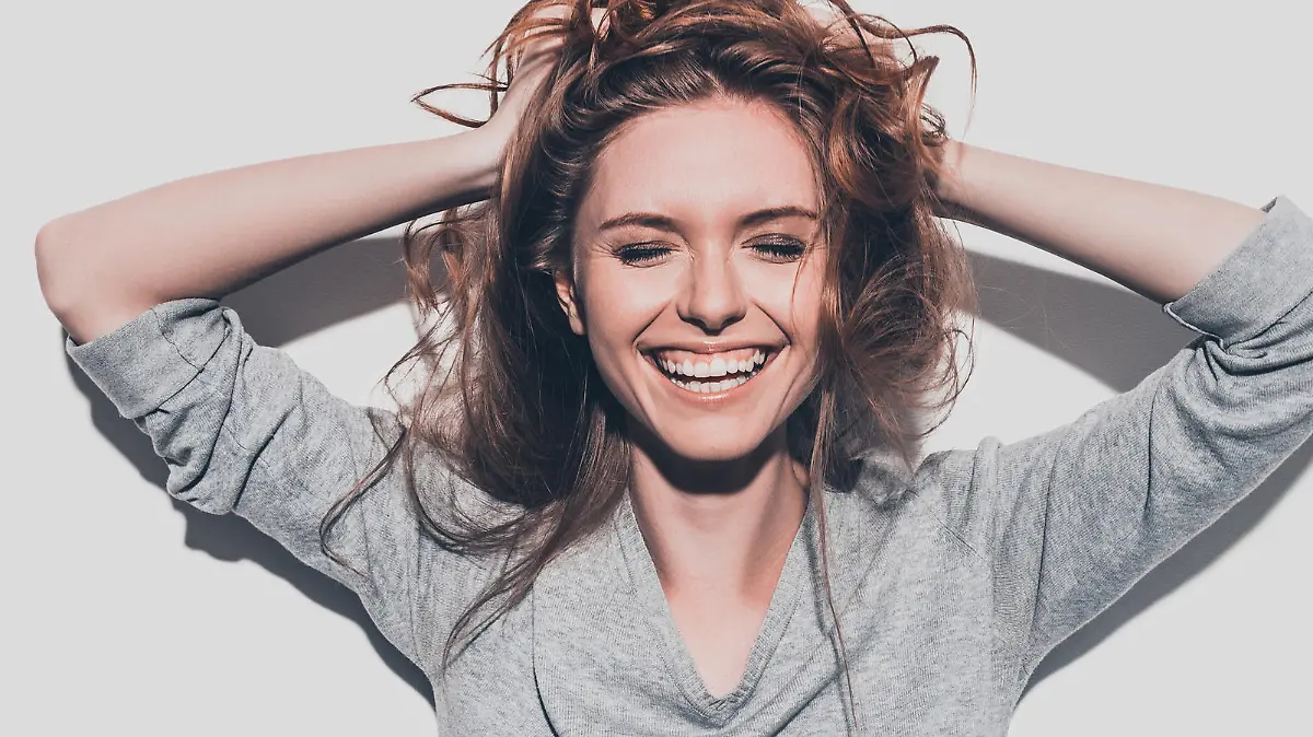Attractive young smiling woman holding hands in hair and keeping eyes closed while standing against grey background