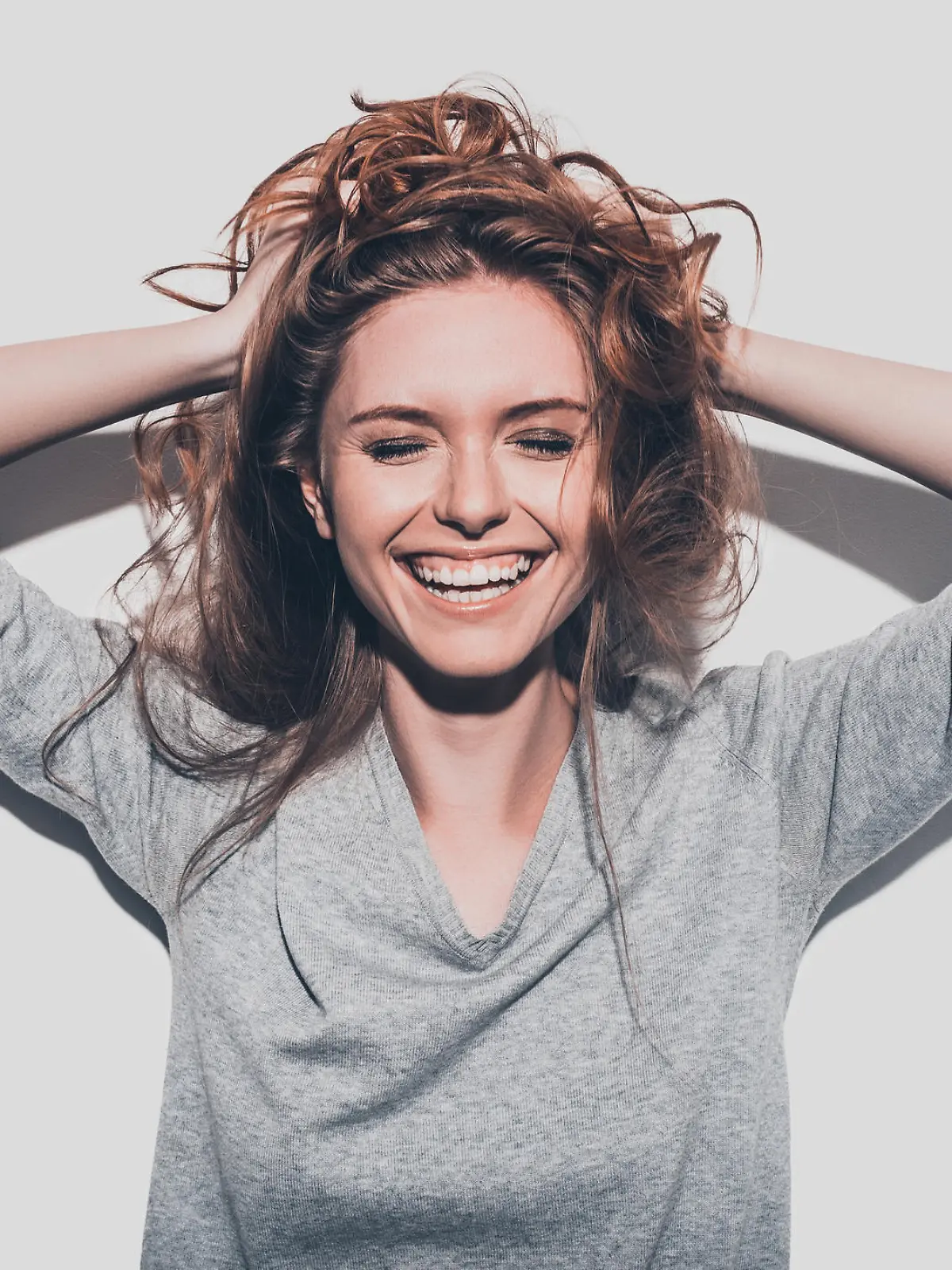 Attractive young smiling woman holding hands in hair and keeping eyes closed while standing against grey background