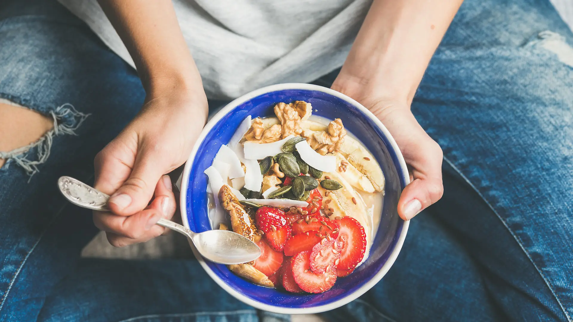 Eating healthy breakfast bowl. Yogurt, granola, seeds, fresh and dry fruits and honey in blue ceramic bowl in woman' s hands. Clean eating, dieting, detox, vegetarian food concept