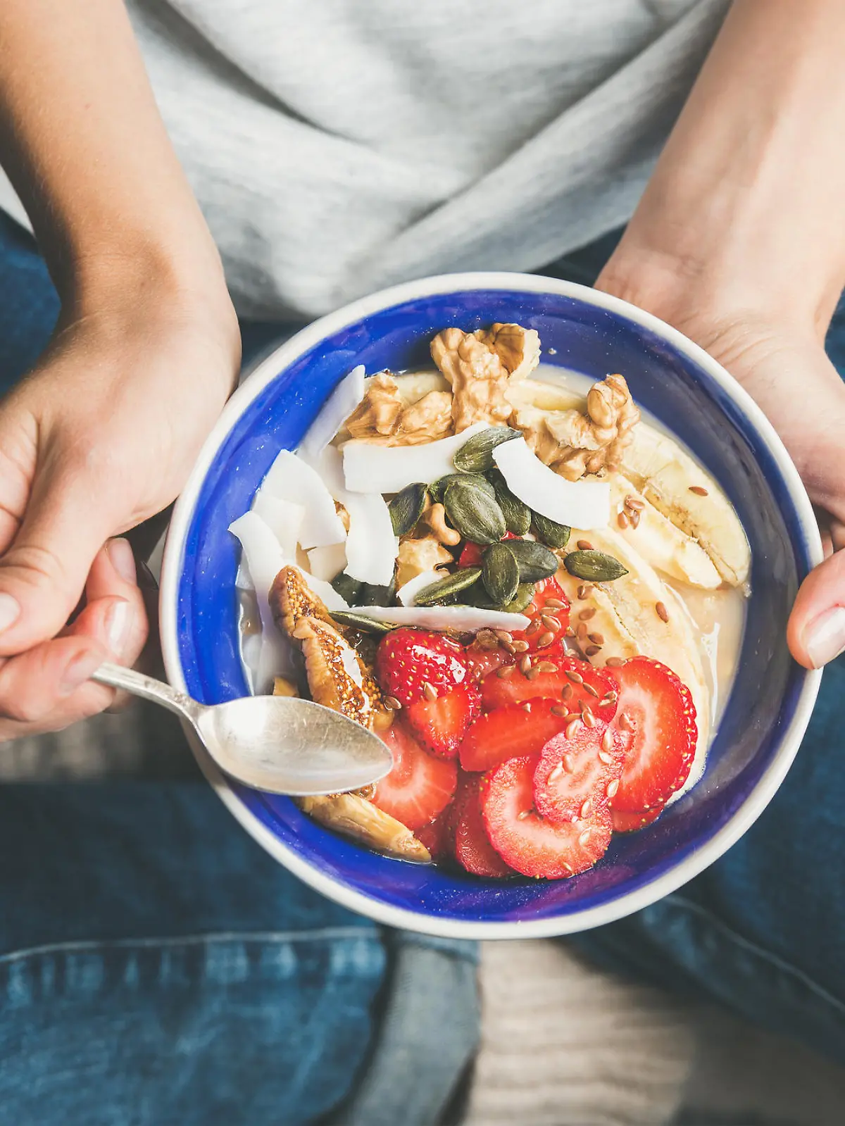 Eating healthy breakfast bowl. Yogurt, granola, seeds, fresh and dry fruits and honey in blue ceramic bowl in woman' s hands. Clean eating, dieting, detox, vegetarian food concept