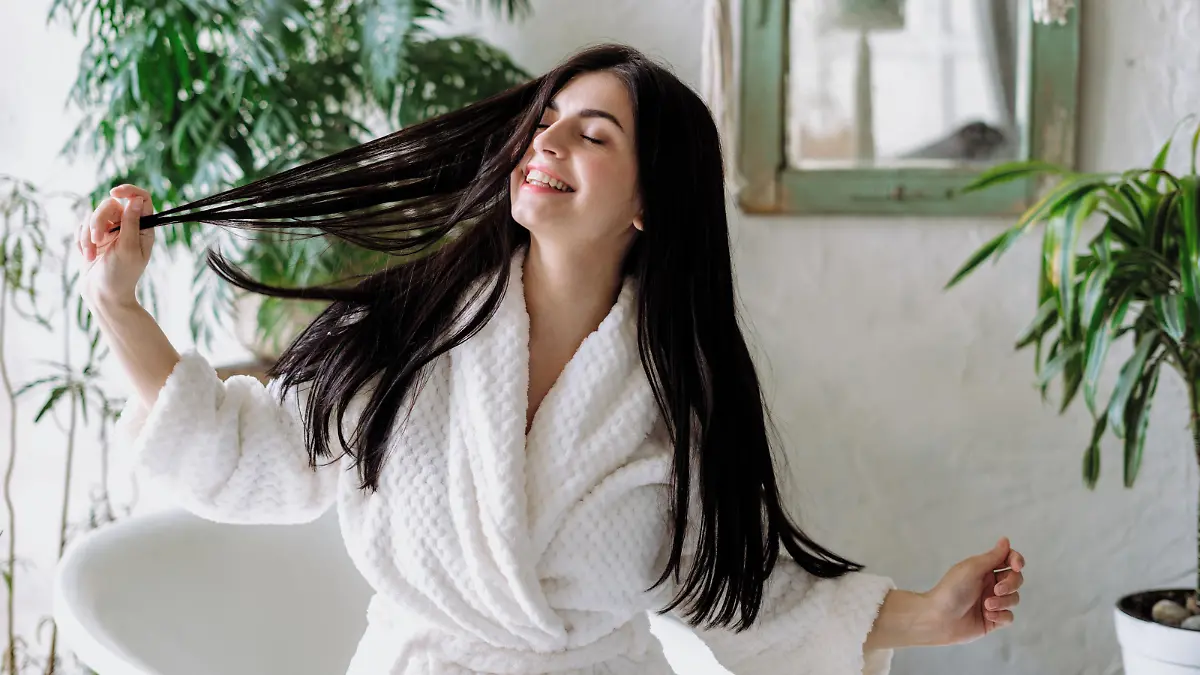 Carefree and happy young adult girl shaking head with healthy floating hair, standing in bathrobe and spending morning at bathroom. Woman having fun at home after daily routine procedure