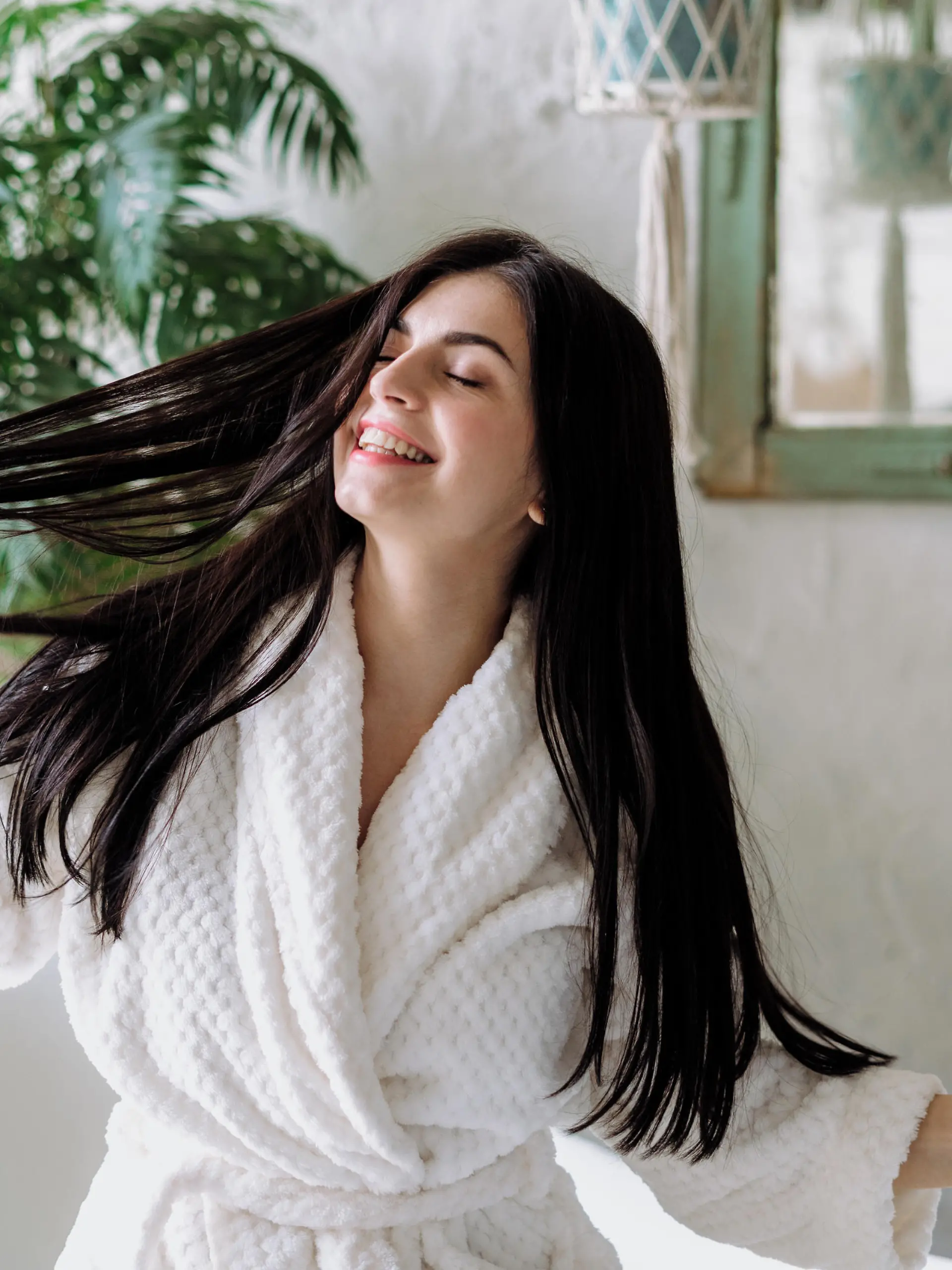 Carefree and happy young adult girl shaking head with healthy floating hair, standing in bathrobe and spending morning at bathroom. Woman having fun at home after daily routine procedure
