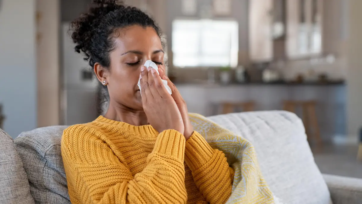 Portrait of young black woman sneezing in to tissue at home. Sick african woman wrapped in blanket sitting on sofa blowing her nose at home. Ill girl sneezing with runny nose in winter.