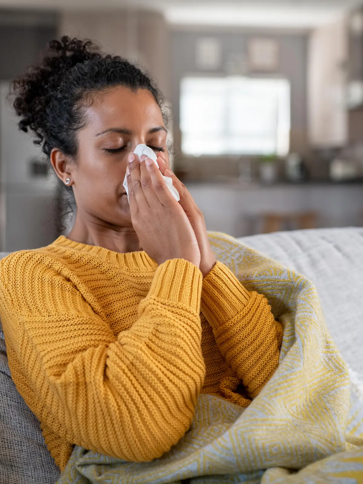 Portrait of young black woman sneezing in to tissue at home. Sick african woman wrapped in blanket sitting on sofa blowing her nose at home. Ill girl sneezing with runny nose in winter.