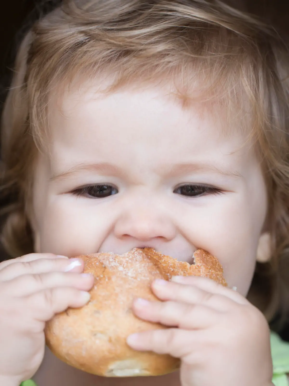 Hungry little cute baby boy with blonde curly hair holding and biting fresh delicious bread roll on black backdroung, horizontal picture