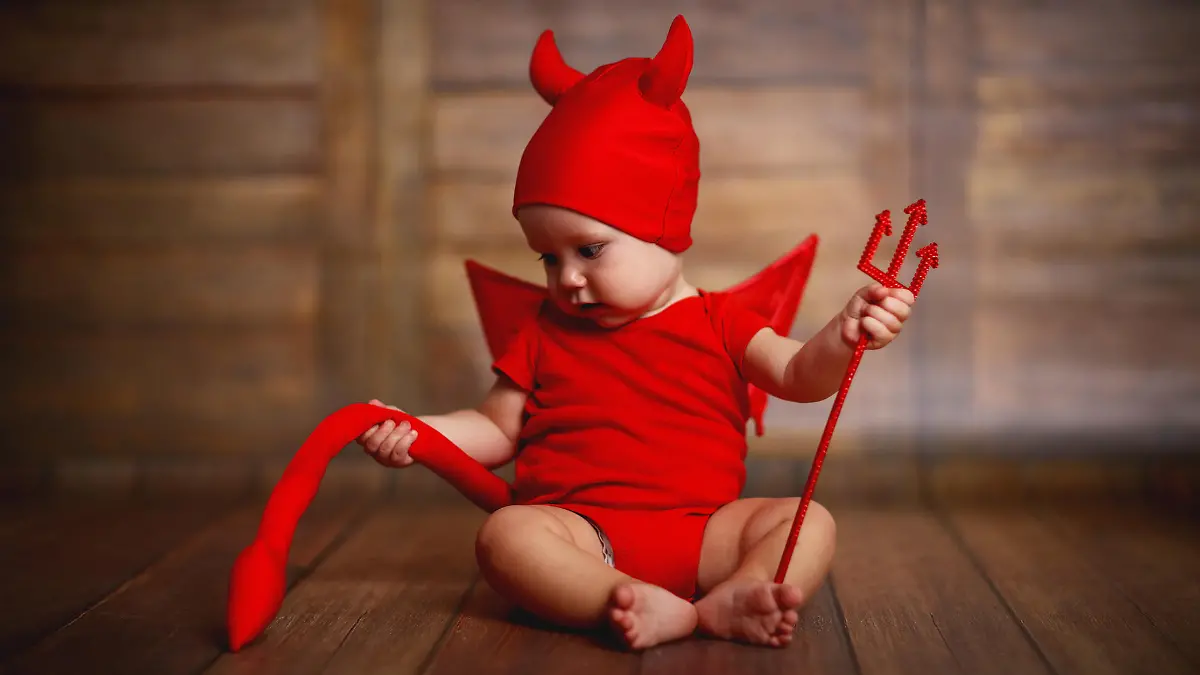 funny baby in devil halloween costume with horns and trident on a dark wooden background