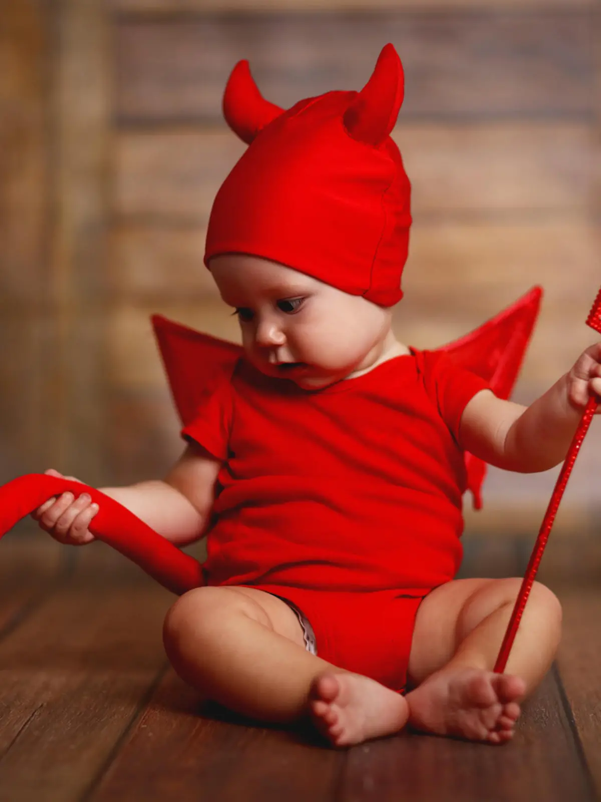 funny baby in devil halloween costume with horns and trident on a dark wooden background