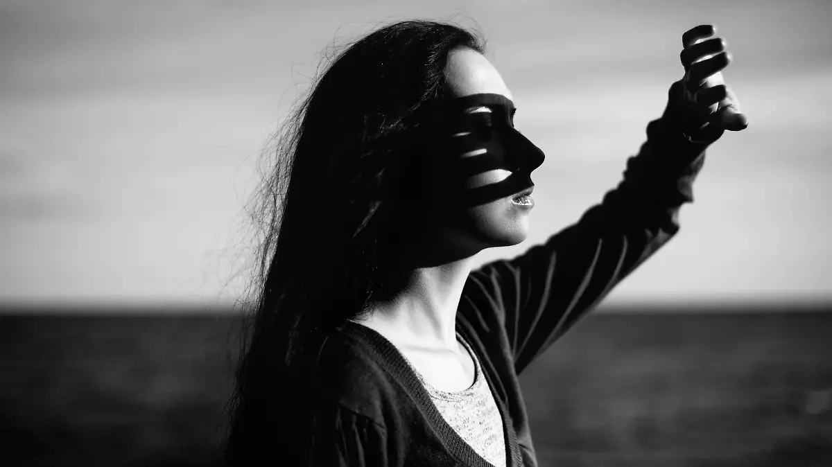 Real young girl in casual wear illuminated by the setting sun near the sea with her hand covering her face from sun light. black and white