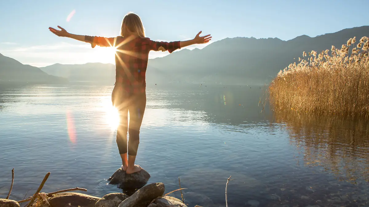 Young cheerful woman by the lake enjoying nature. Arms outstretched for positive emotion. 