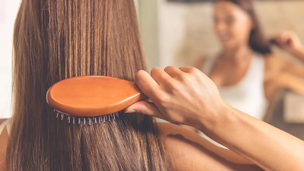 Back view of beautiful young woman in white undershirt combing her hair and smiling while looking into the mirror in bathroom