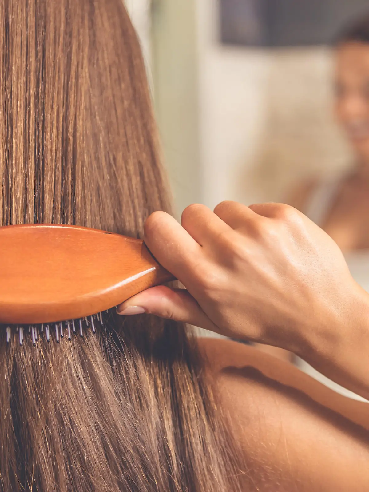 Back view of beautiful young woman in white undershirt combing her hair and smiling while looking into the mirror in bathroom