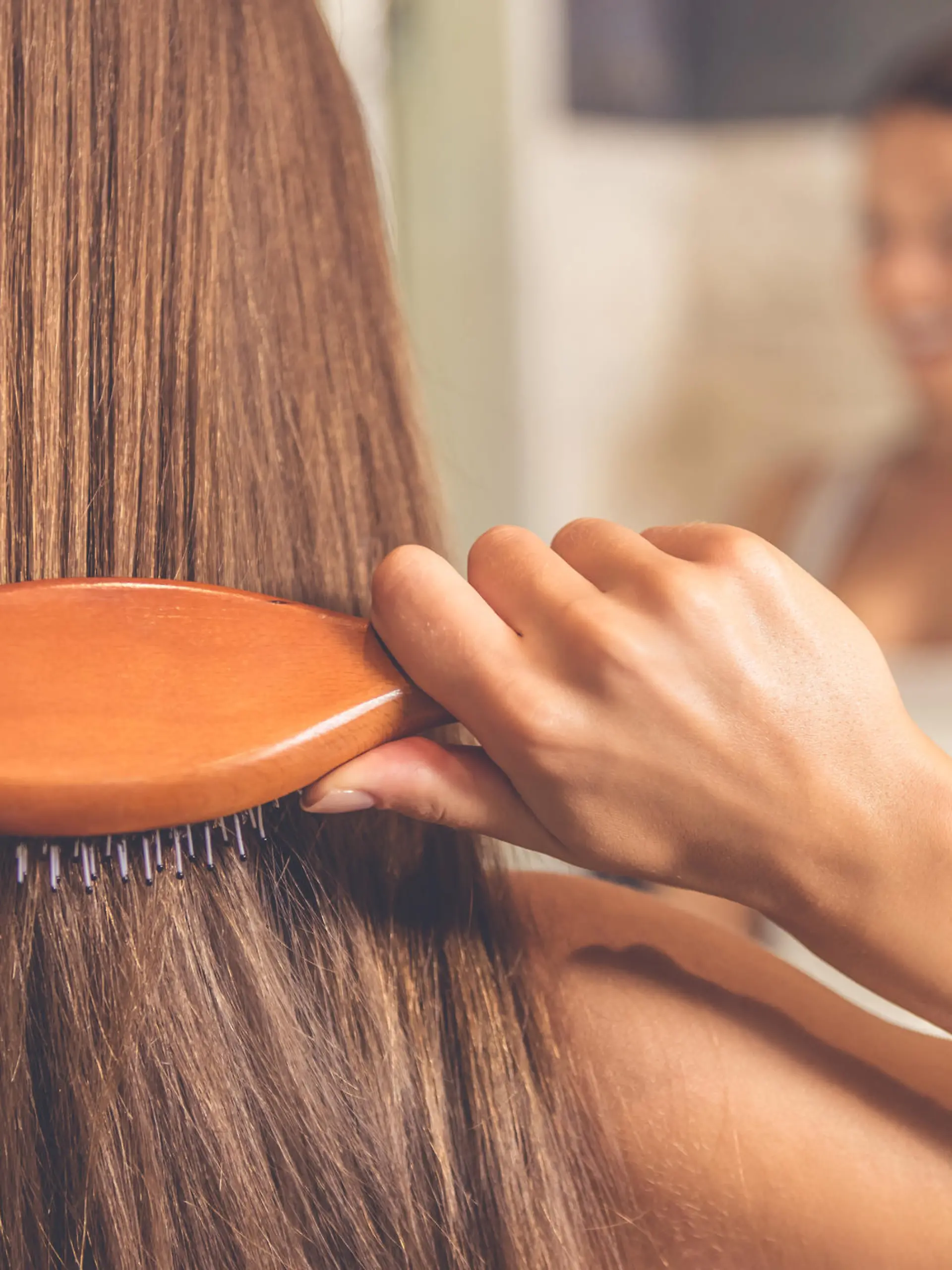 Back view of beautiful young woman in white undershirt combing her hair and smiling while looking into the mirror in bathroom