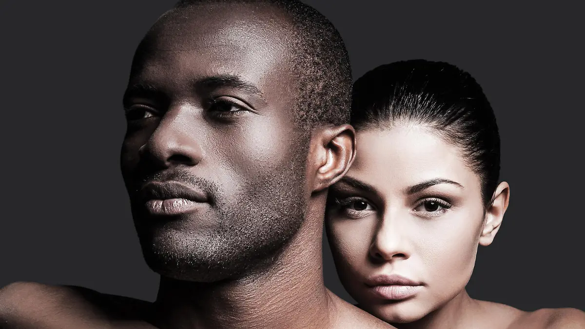 Beautiful Caucasian woman bonding to handsome African man and looking at camera while both standing against grey background