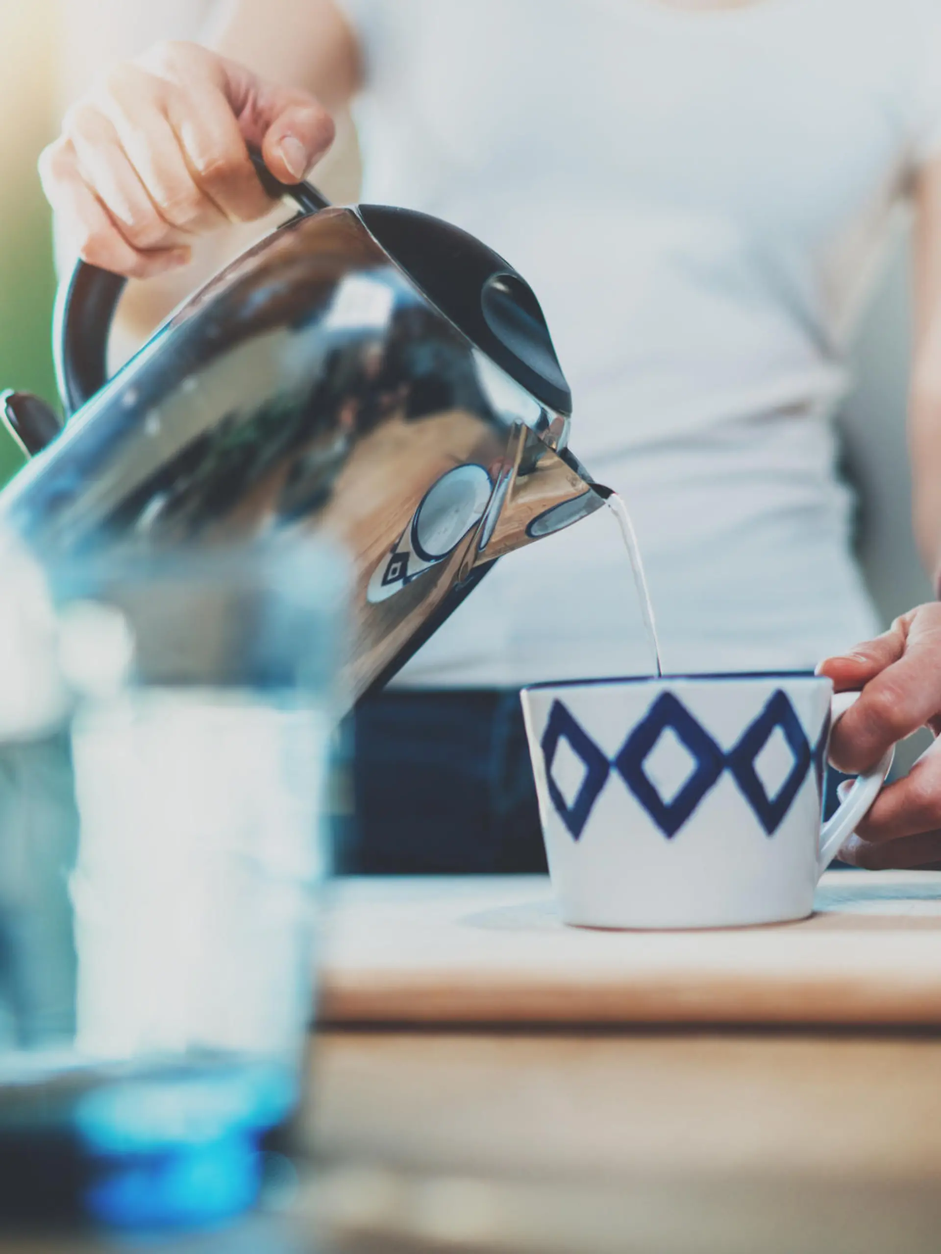 Young woman using kettle for make tea or black coffee on kitchen at room interior background.Women's hands pour water from a teapot into a cup. Blurred, flares effect