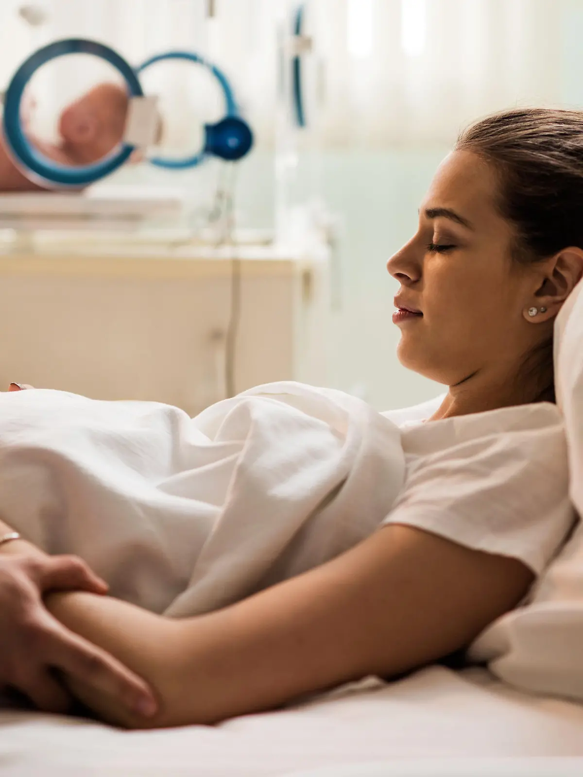 Young woman lying in hospital bed with her eyes closed after childbirth while unrecognizable person is consoling her.
