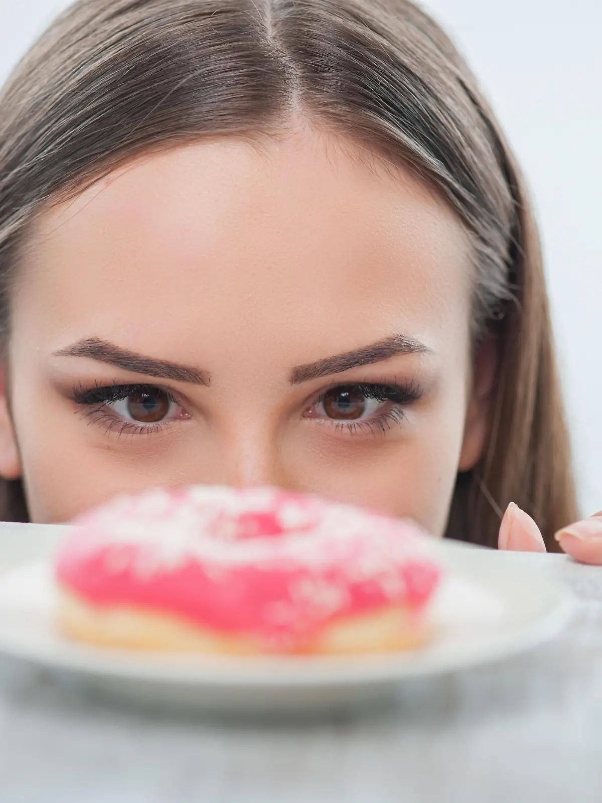 Beautiful girl is looking at unhealthy donut with appetite. It is situated on a table. Isolated on a white background
