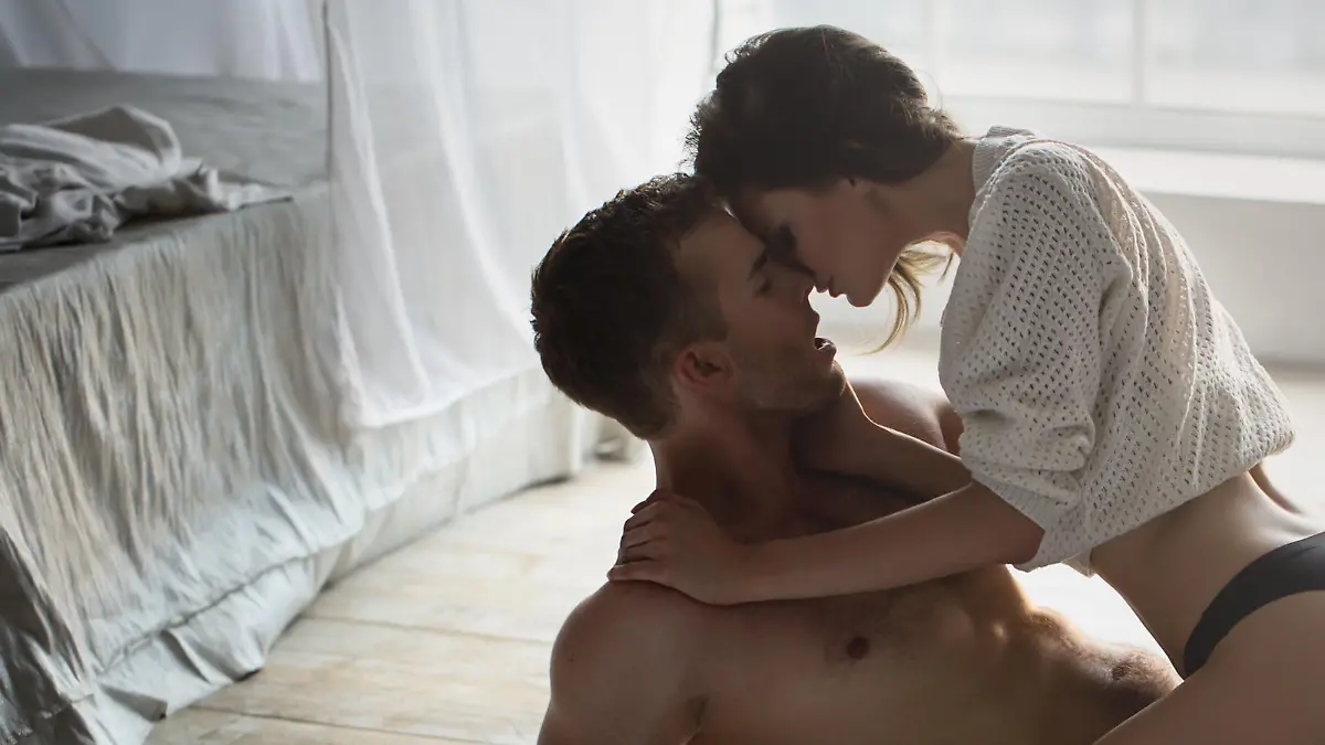 Passionate couple kissing, boy and girl sitting on wooden floor near the rumpled bed opposite the window.