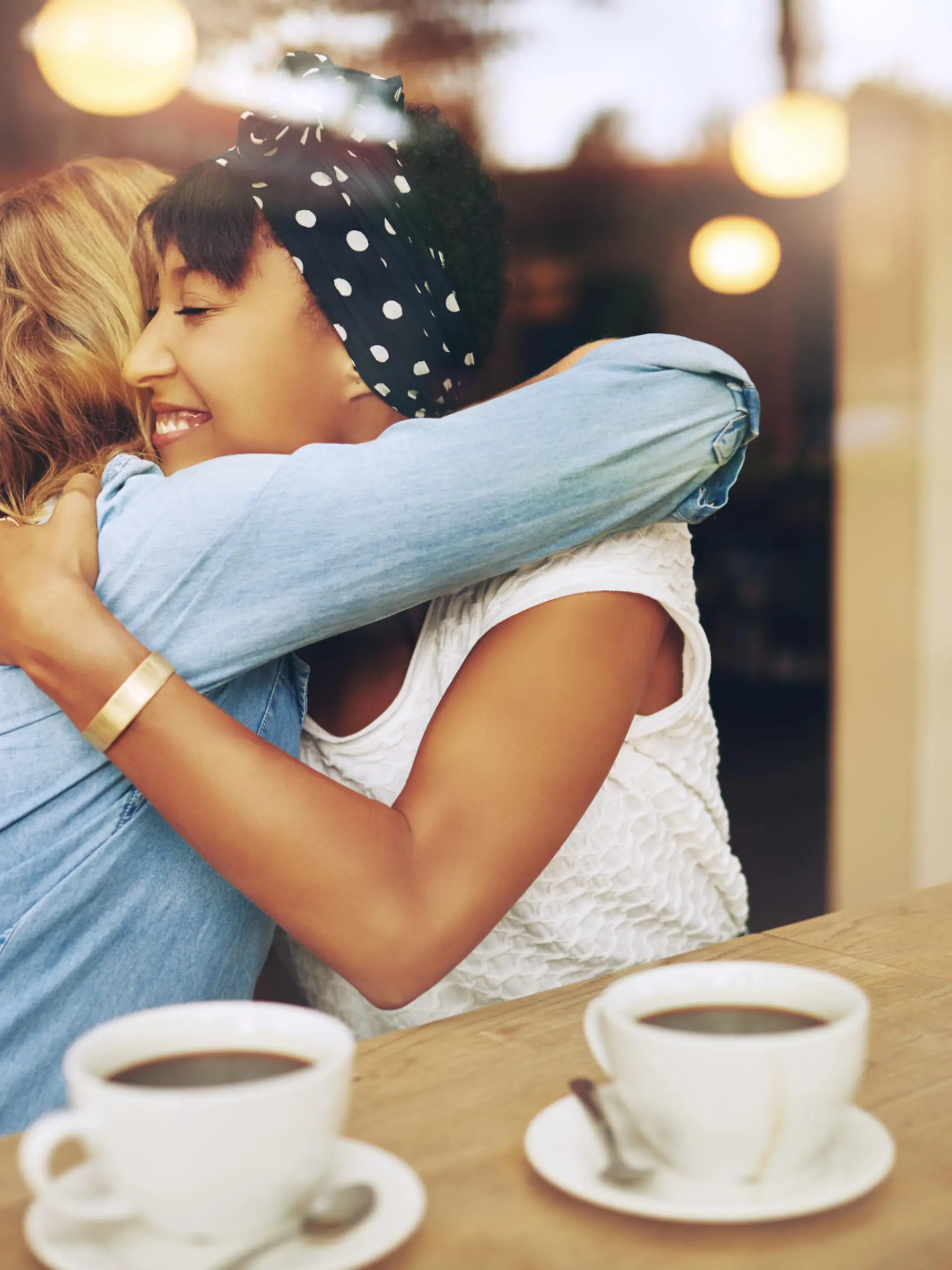 Two multi ethnic affectionate girl friends embracing as they sit in a coffee shop enjoying a cup of coffee together