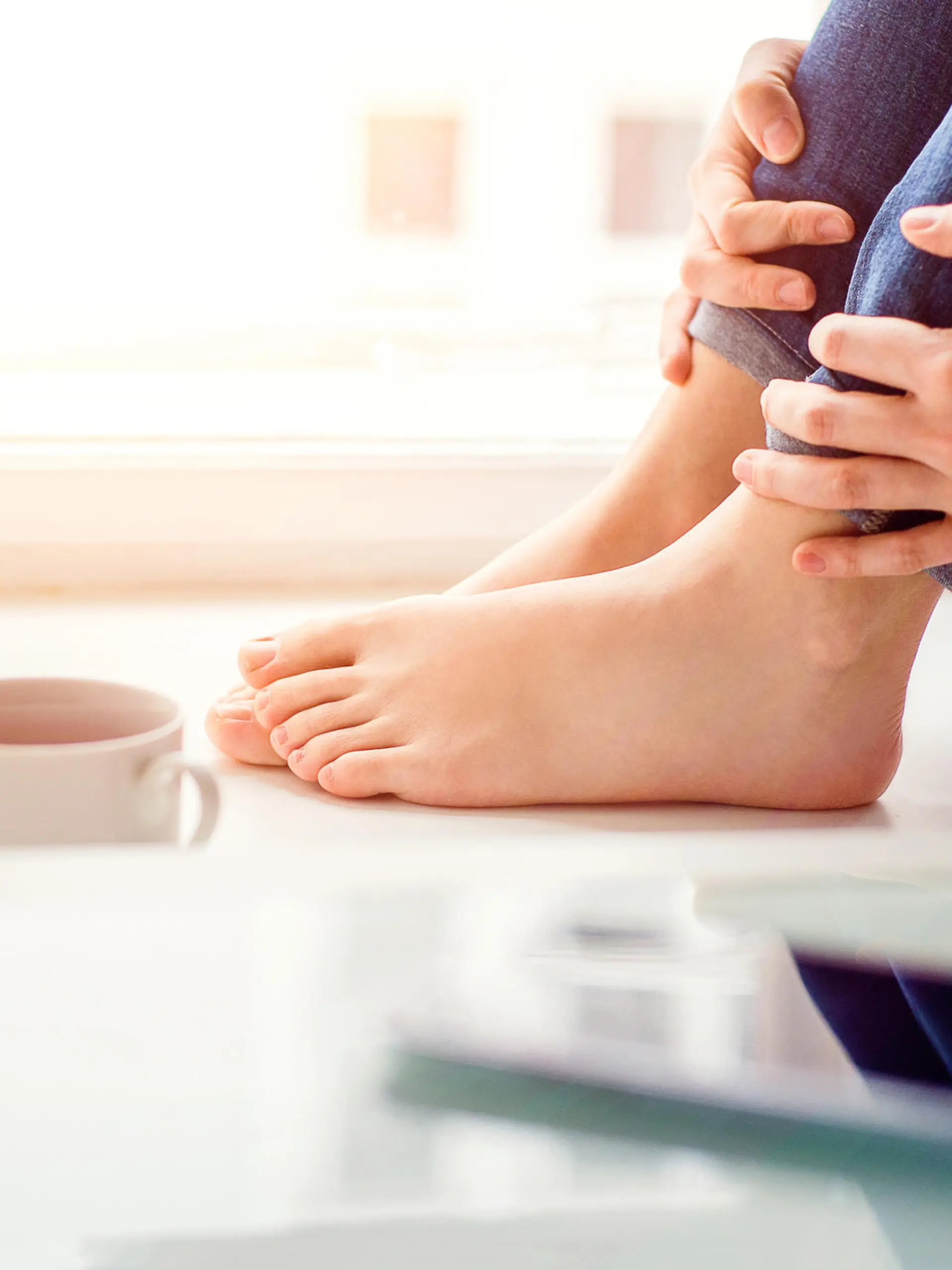 Feet of unrecognizable woman sitting on window sill with smart phone and cup of coffee