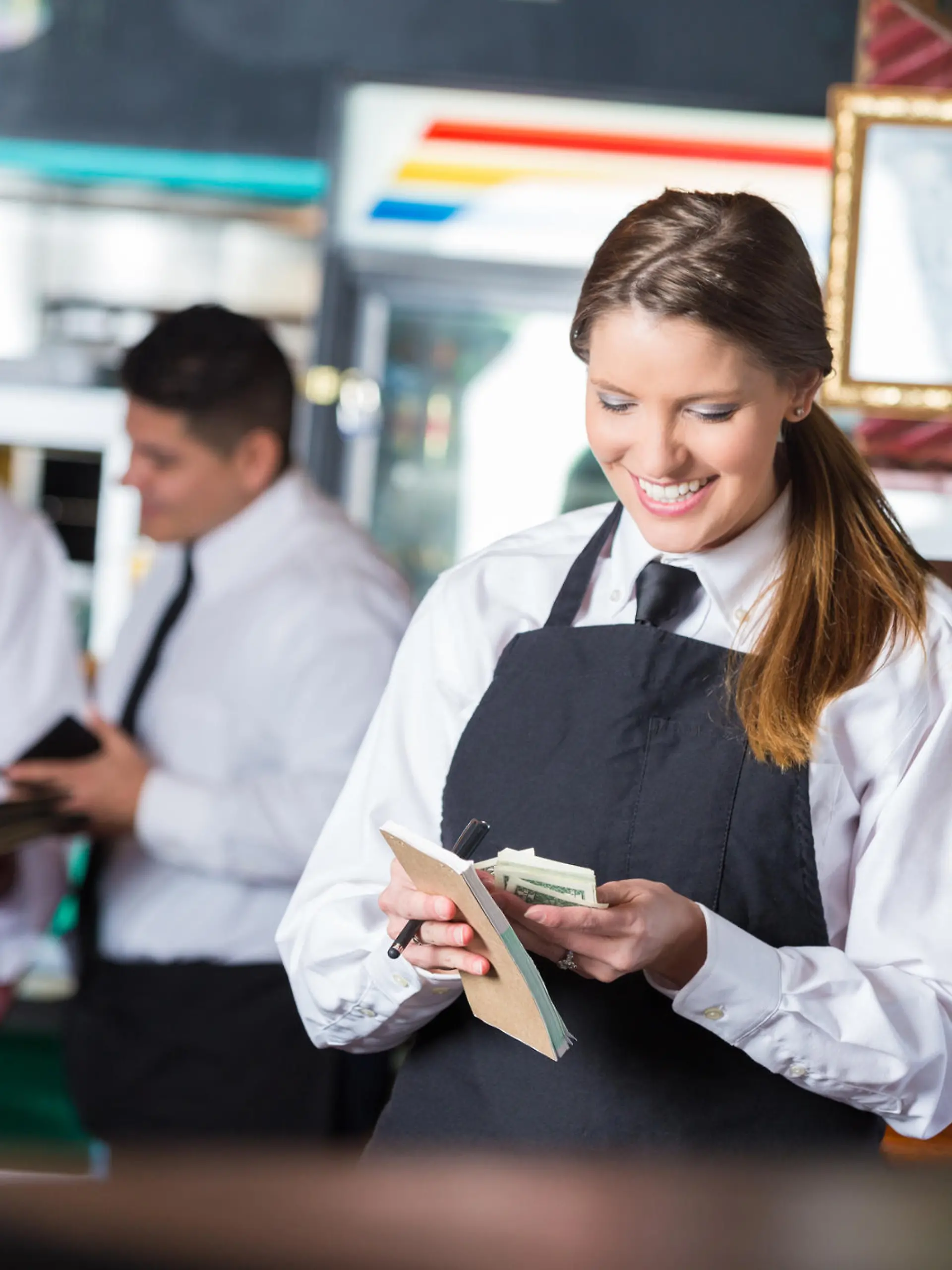 Pretty mid adult Caucasian woman is standing in front of restaurant commercial kitchen. She is wearing a white shirt with a black tie and apron, and is a professional waitress. She's smiling while looking down and counting cash tips after working long shift. Chef and restaurant manager are standing in background of casual dining restaurant. 