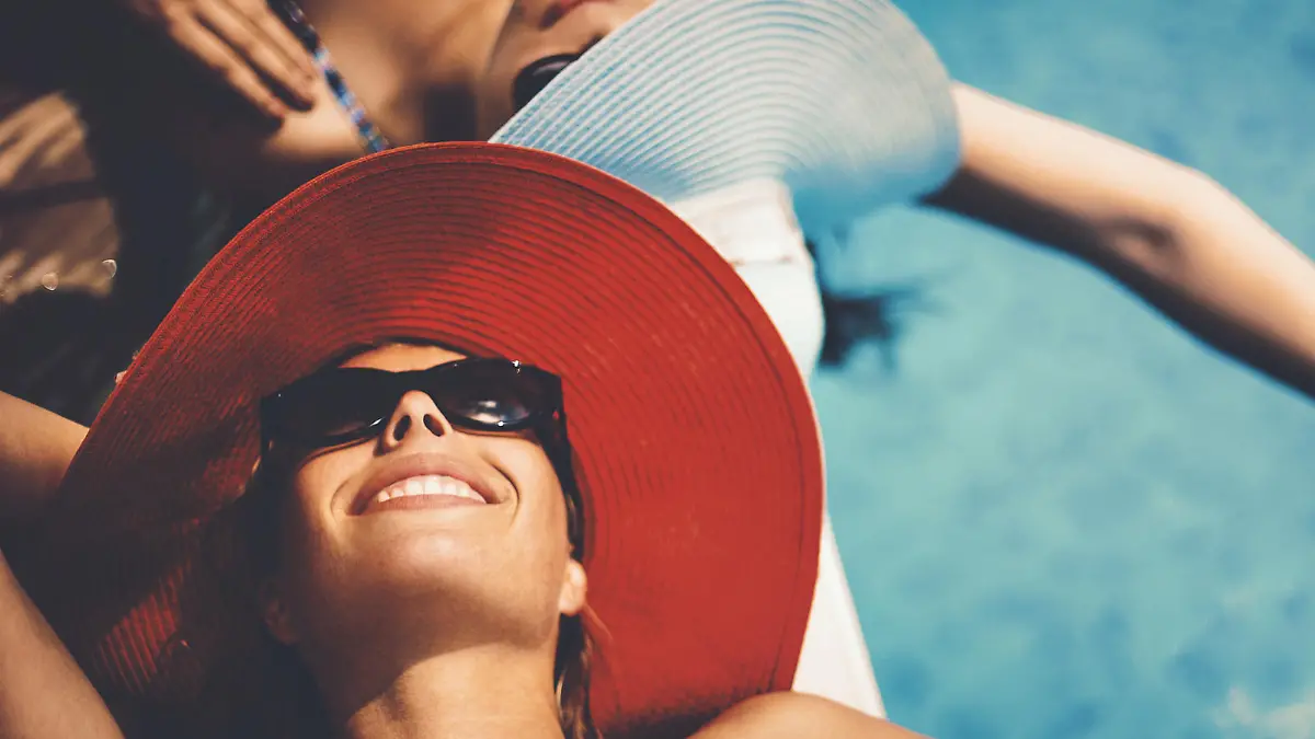 Closeup of two mid 20's women sunbathing by a pool side on hot summer day. Both wearing hats and sunglasses and holding hands above water. Closeup, top view.
