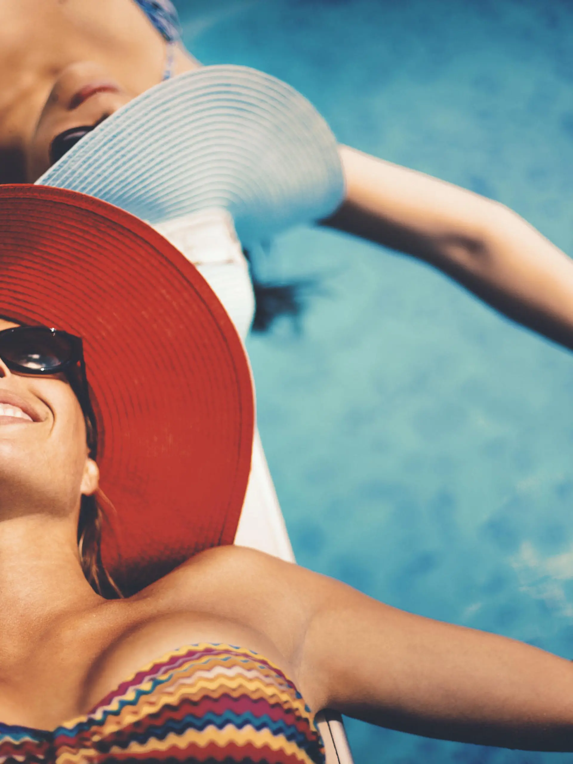 Closeup of two mid 20's women sunbathing by a pool side on hot summer day. Both wearing hats and sunglasses and holding hands above water. Closeup, top view.