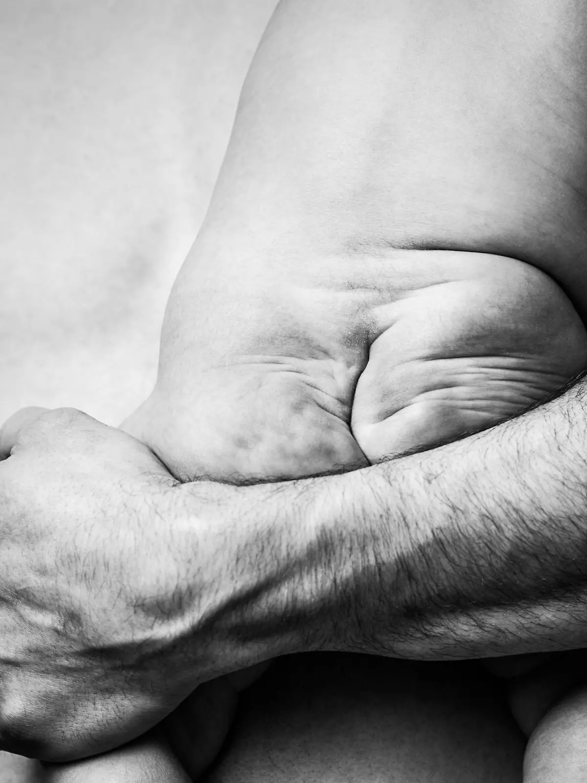 A cute low key image of a squishy babies butt in all its naked glory.  A dad holds the baby close to his bare chest.  Black and white, horizontal image.