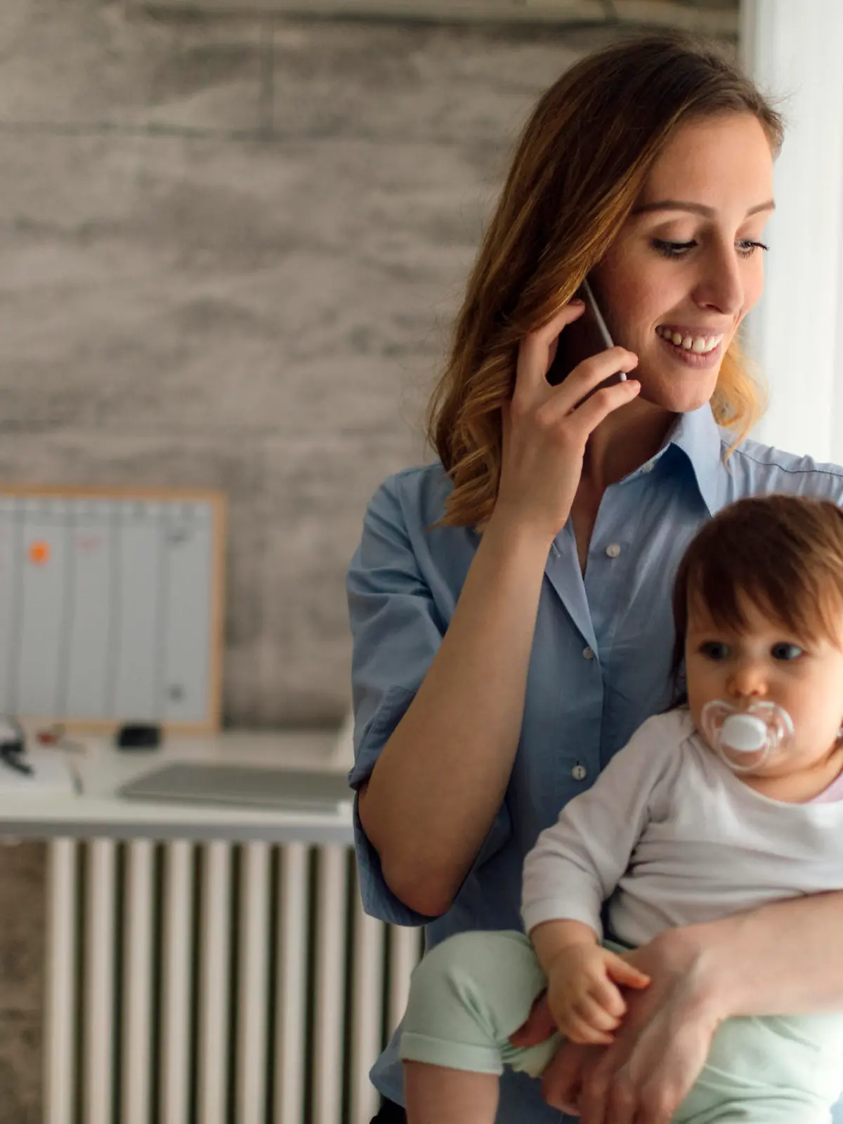Mother holding smart phone and talking while carrying her cute baby girl. She is happy and cheerful. Mother is working from home. Baby has baby pacifier.