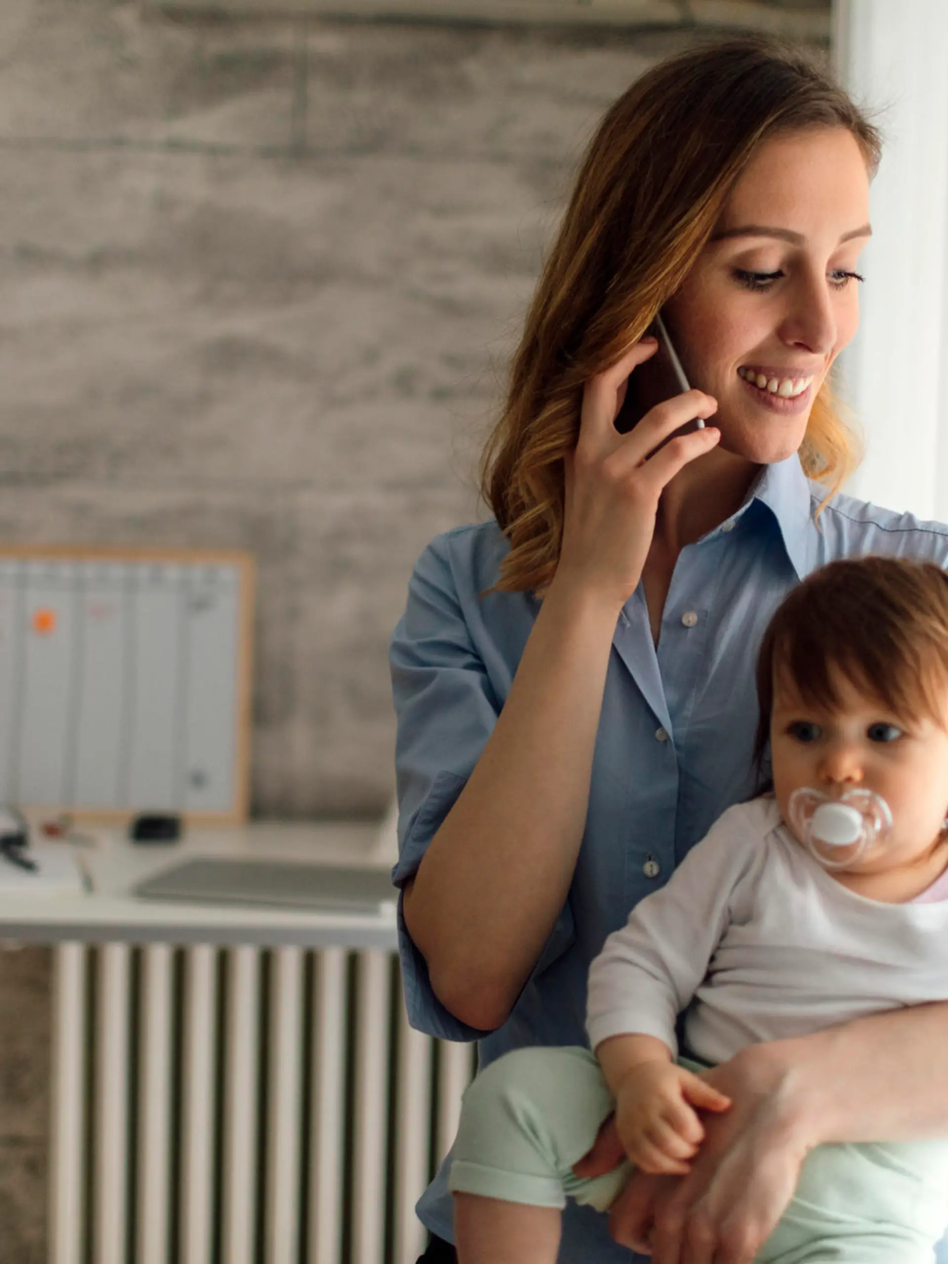 Mother holding smart phone and talking while carrying her cute baby girl. She is happy and cheerful. Mother is working from home. Baby has baby pacifier.
