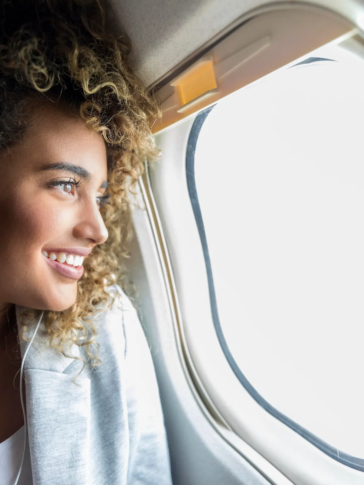 Confident young mixed race woman smiles while looking through window on aircraft. She is wearing earbuds.