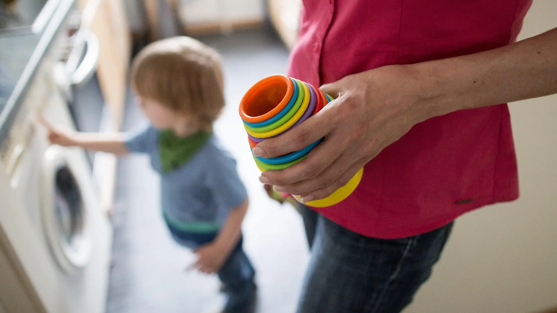 Eine Mutter hält am 01.07.2016 in Dortmund (Nordrhein-Westfalen) Kinderspielzeug in der Hand, während ein kleiner Junge an einer Waschmaschine spielt. Den bundesweit rund 2,3 Millionen Kindern von Alleinerziehenden droht deutlich häufiger Armut als ihren Altersgenossen in Paarfamilien. Foto: Marcel Kusch/dpa (zu dpa "Studie: Immer mehr Kinder von Alleinerziehenden in der Armutsfalle" vom 06.07.2016) +++(c) dpa - Bildfunk+++