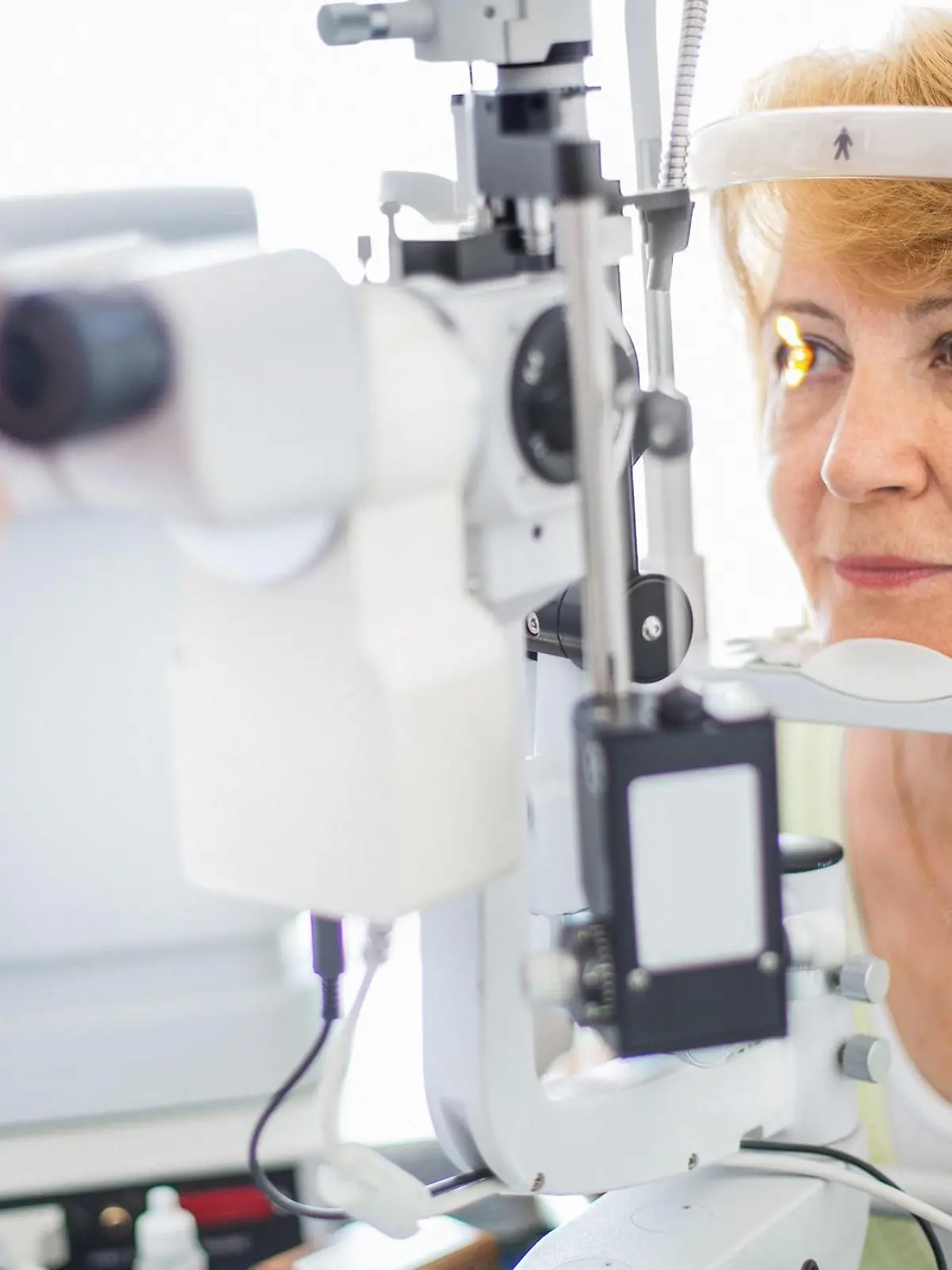 Senior caucasian woman having her eyes examined at the optician.Her head is placed in phoropter apparatus while middle aged male doctor is examining her retina. The woman has mid length yellow brown hair and wearing light breen blouse.