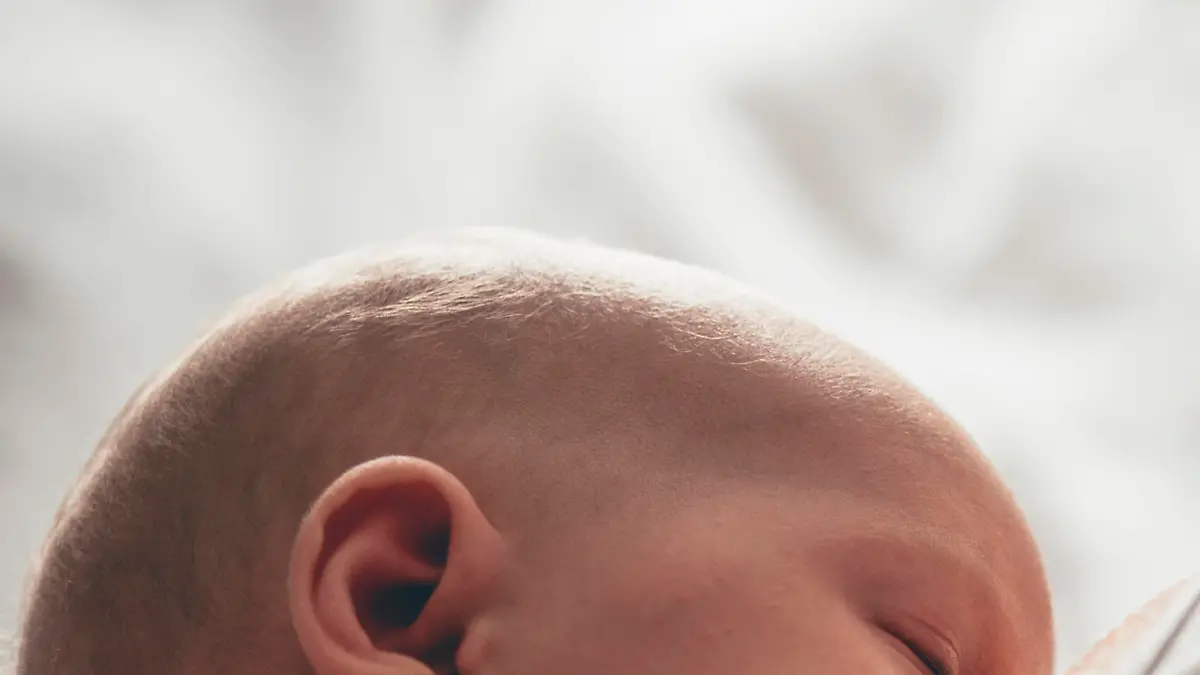 A newborn baby sleeps in her mother's arms while breastfeeding from her mother. The baby's face in in focus as the mother is unrecognisable.
