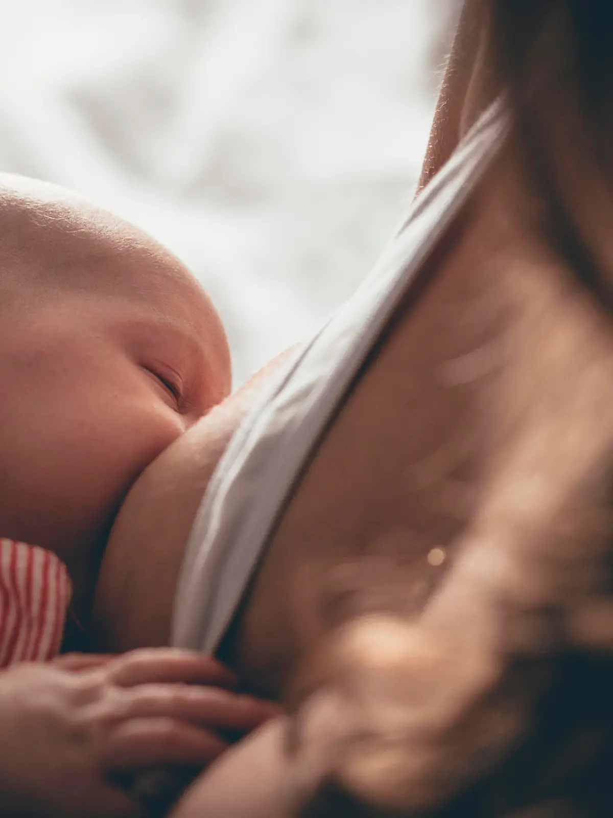 A newborn baby sleeps in her mother's arms while breastfeeding from her mother. The baby's face in in focus as the mother is unrecognisable.