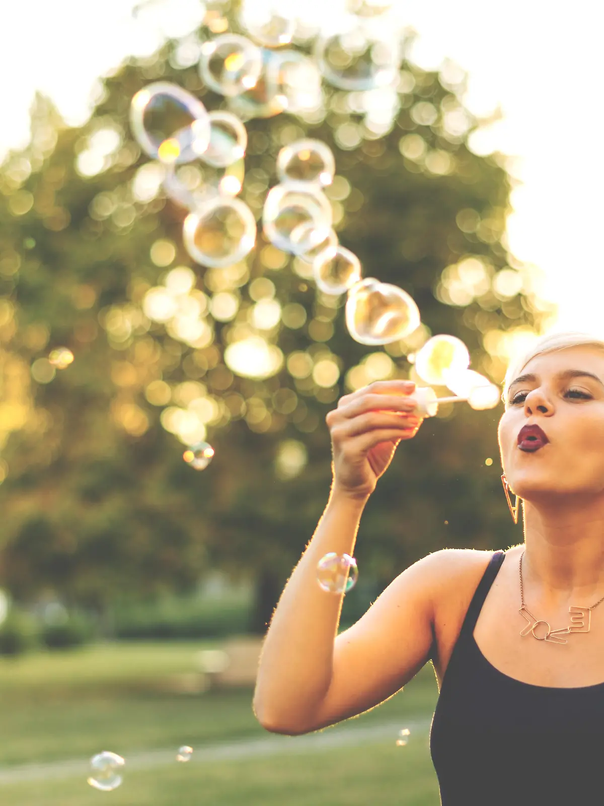 Portrait of modern hipster blonde woman, 20-25 years old in park, relaxing, blowing bubbles and having leisure time on her own. Weekend activities in summer.