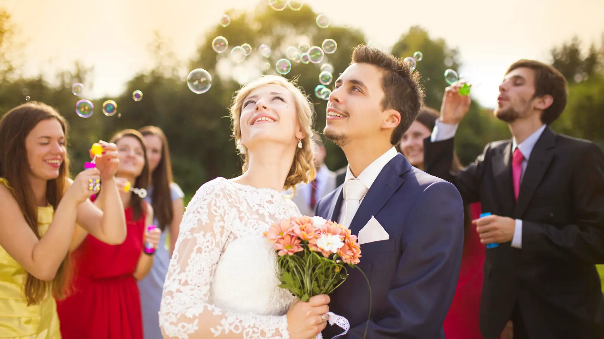 Young newlyweds enjoying romantic moment together at wedding reception outside, wedding guests in background blowing bubbles