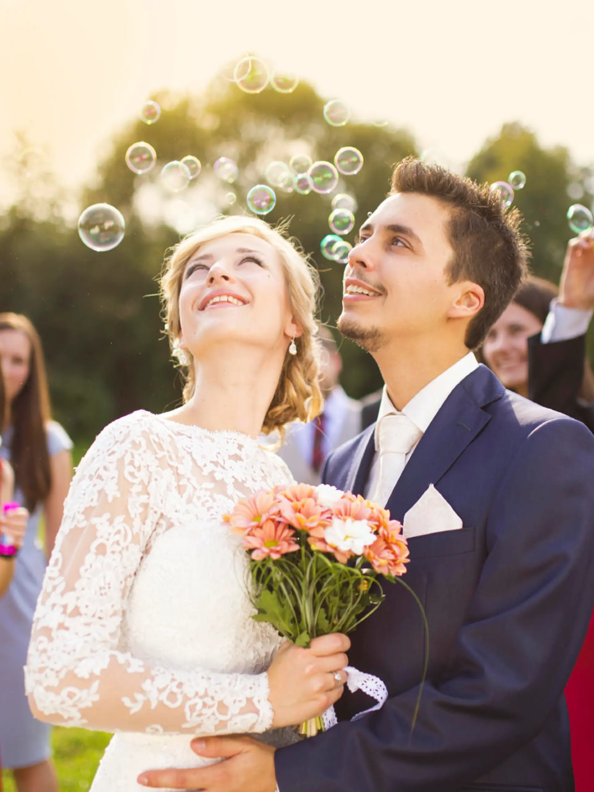 Young newlyweds enjoying romantic moment together at wedding reception outside, wedding guests in background blowing bubbles